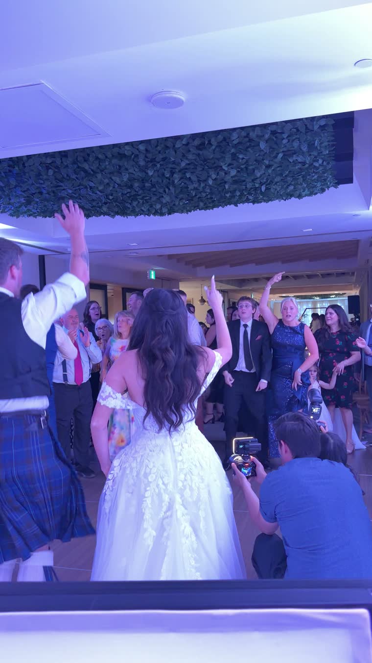 A bride with her arm raised faces a cheering crowd on the dance floor at Andaz Ottawa ByWard Market